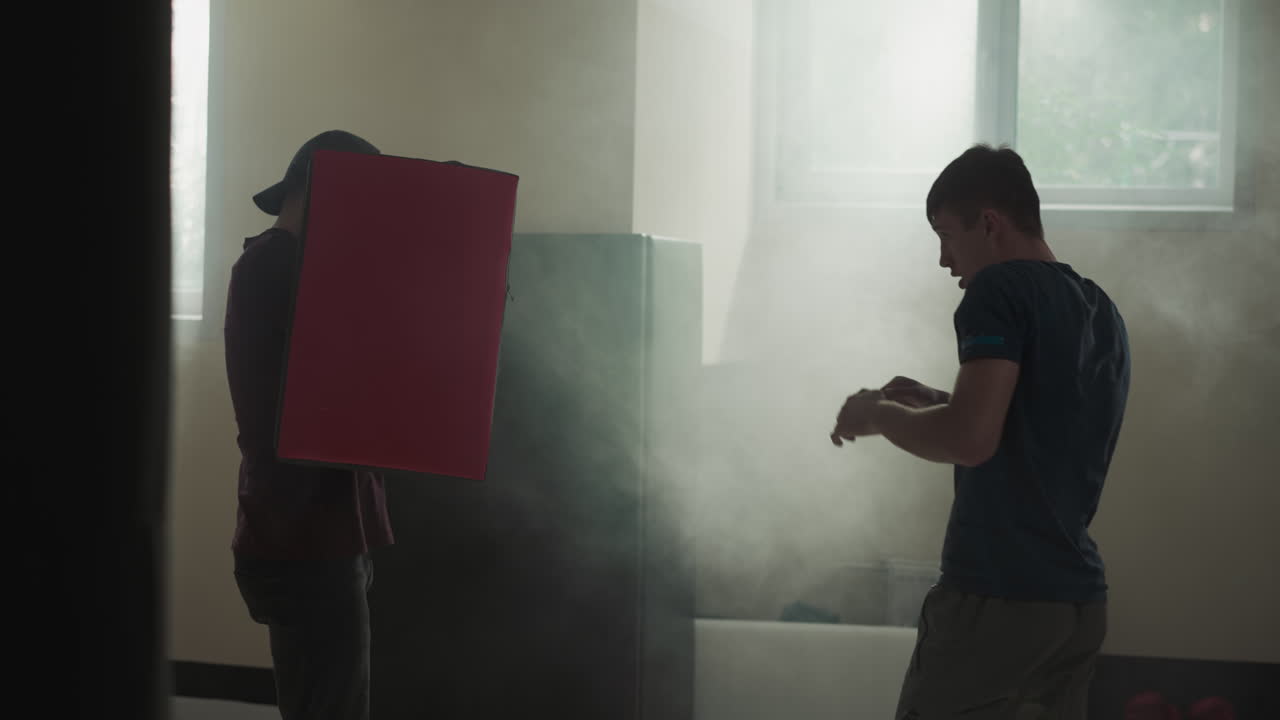 활기찬 남자 실루은 스포츠 클럽의 지하실에서 맨발로 목표물을 적극적으로 타격합니다. 남성 트레이너는 어두운 전제에서 성공으로 학생을 이끌 수있는 장비를 들고 있습니다.