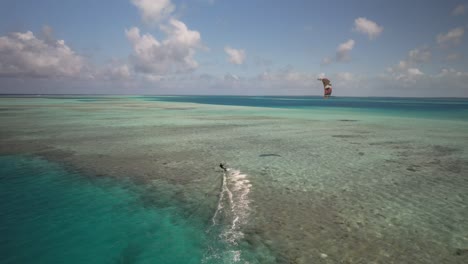 Kitesurfer-Gleitet-über-Klares,-Blaues-Tropisches-Wasser-Mit-Drachen-Am-Himmel,-Kamerafahrt-Per-Drohne-Aus-Der-Luft