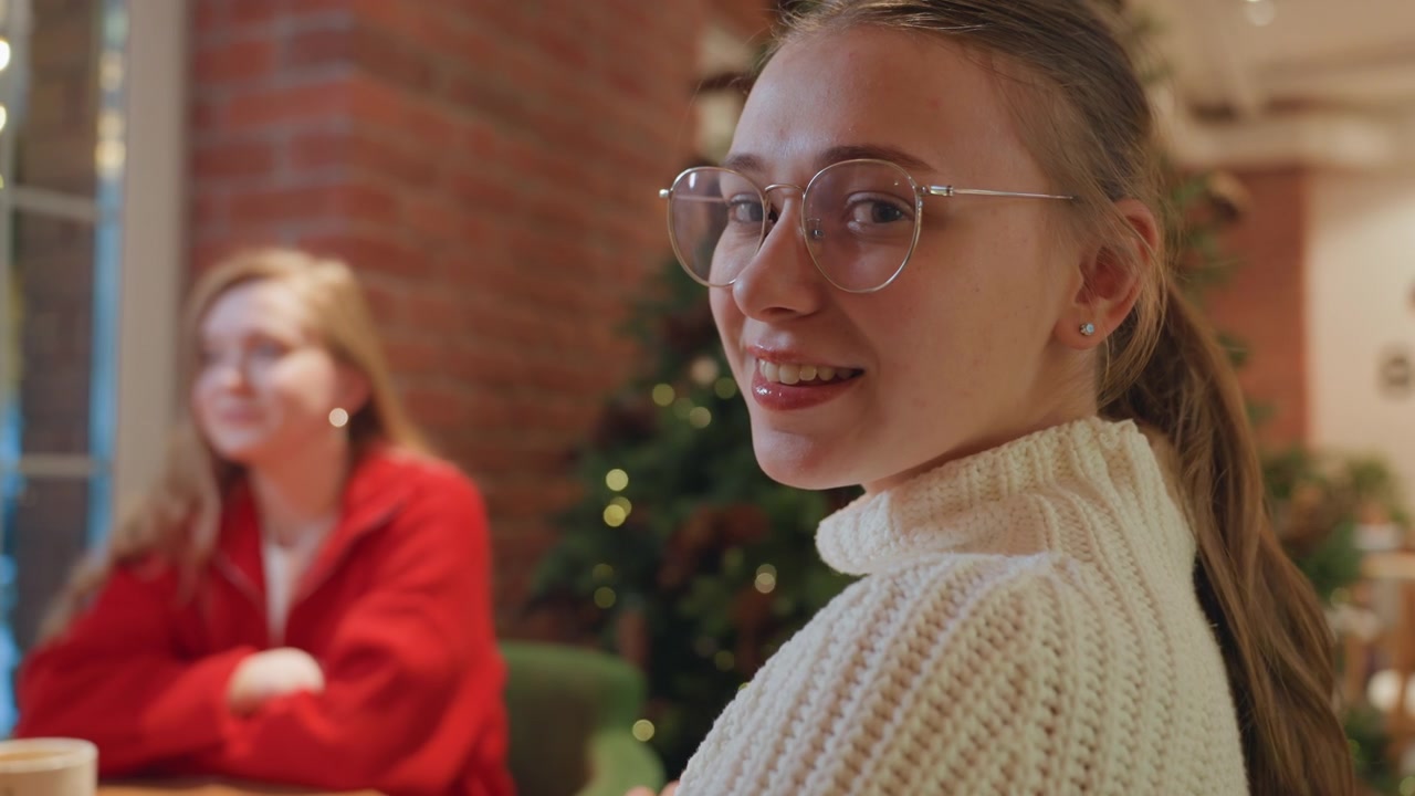 Young Woman Smiles at Camera While Friend Talks in Christmas Cafe