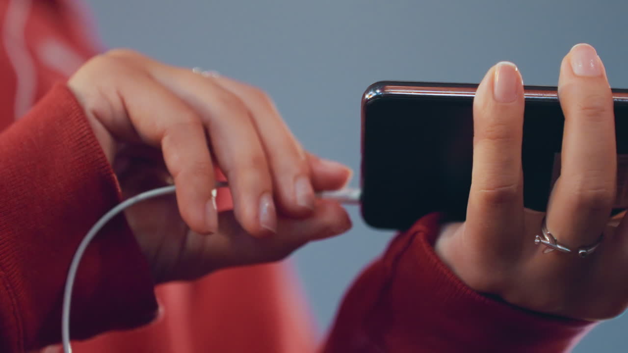 Mani in primo piano che inseriscono auricolari nello smartphone, donna bianca con felpa rossa, anelli d'argento sulle dita, movimenti lenti e deliberati, rituale d'ascolto intimo, illuminazione cinematografica soffusa, profondità di campo ridotta