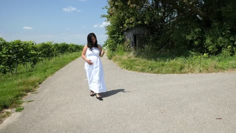 una joven embarazada con un vestido blanco de verano caminando por un camino rural sosteniendo su vientre y flores para una sesión de fotos de maternidad