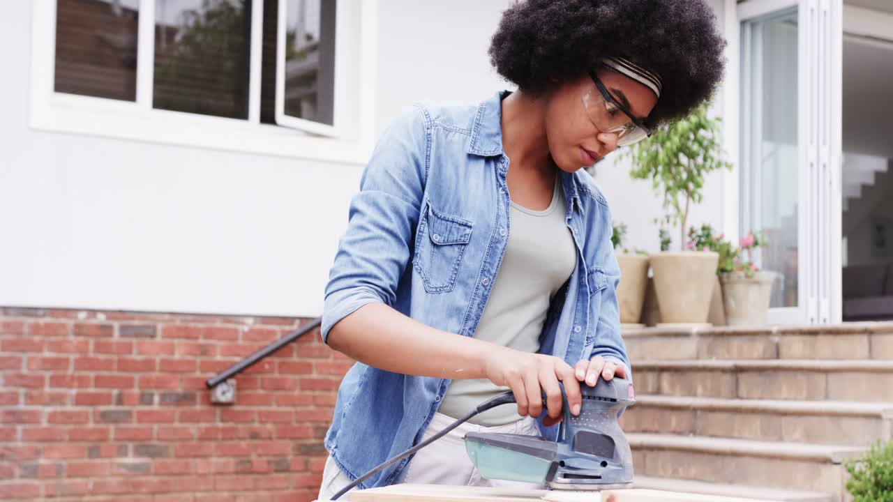 African American woman using corded sander on plank at porch, in denim shirt and goggles, smoothing