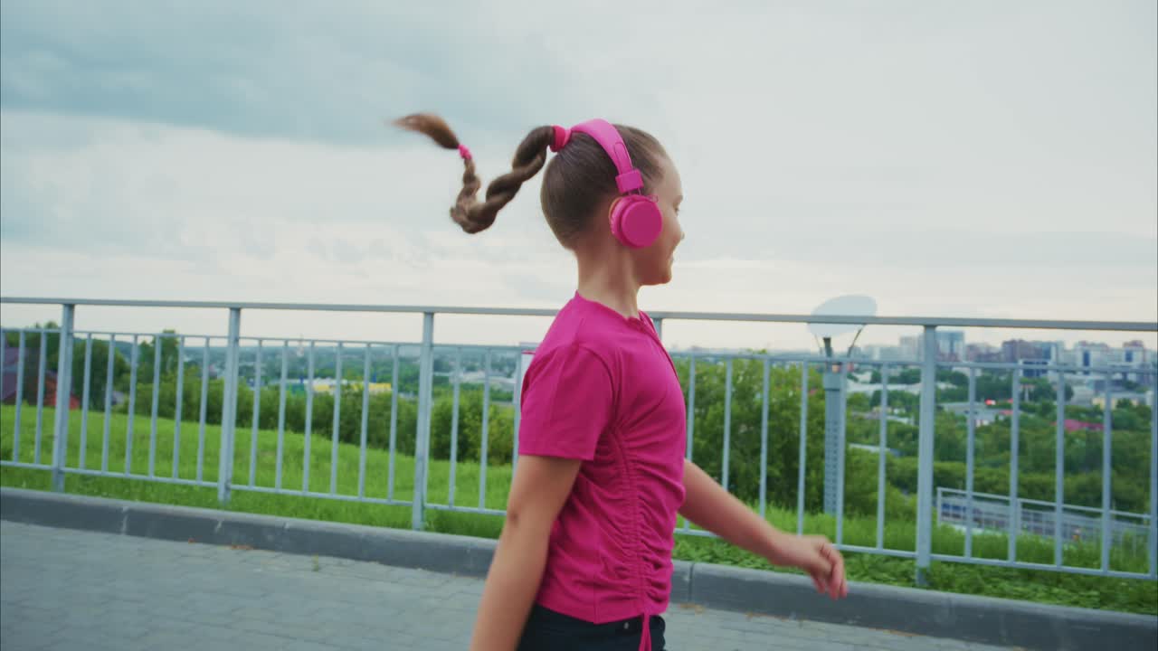 A young girl joyfully walks along a scenic path while listening to music through bright pink headphones, enjoying the beautiful outdoor landscape and fresh air