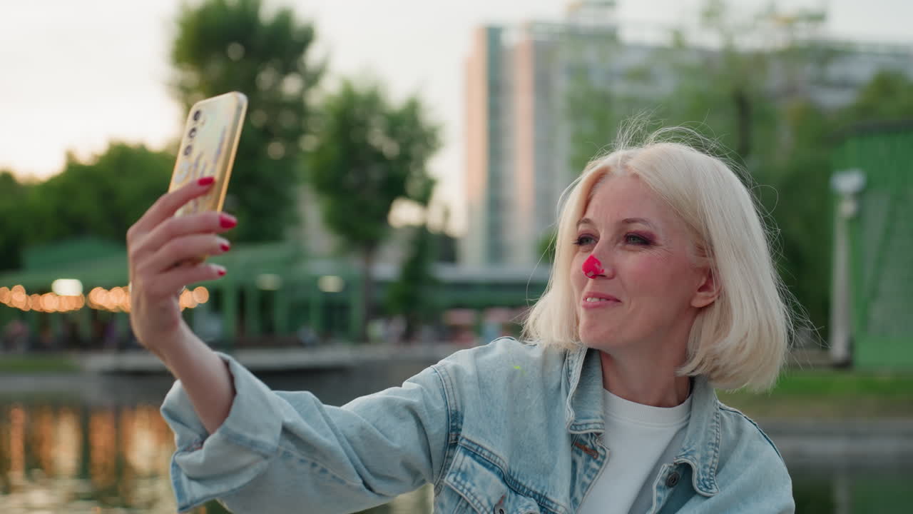 close up smiling mother taking selfie with smartphone by river in green park at golden hour, paint dot on nose, casual denim jacket, blur background with trees water reflecting warm light
