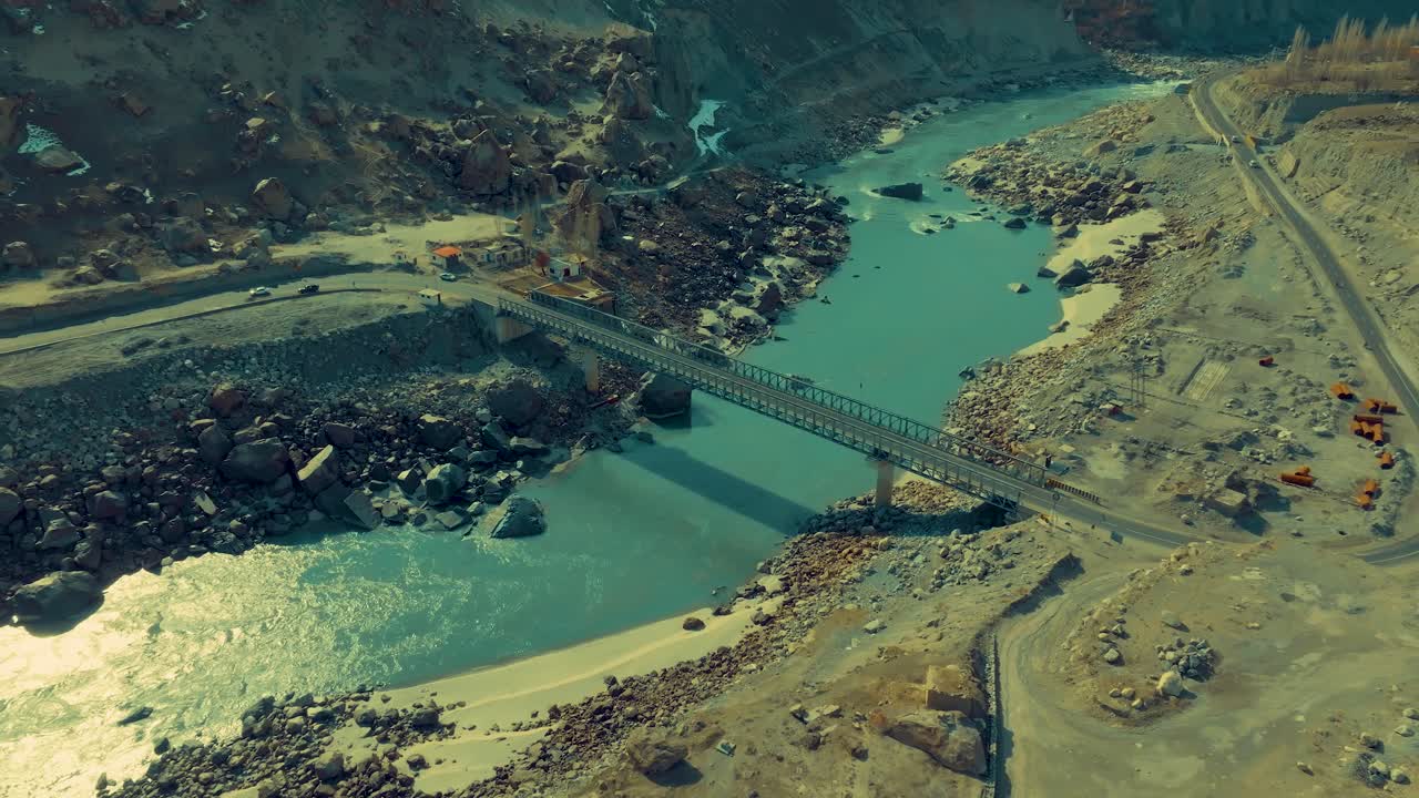 Aerial shot showcasing a modern bridge spanning across a rushing river flanked by rocky terrain and mountains, highlighting the blend of nature and engineering marvel
