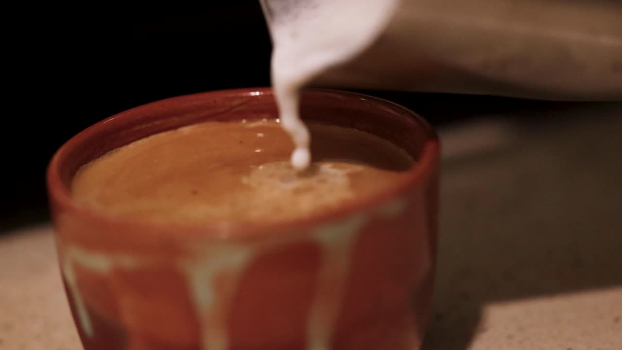 Close-up of milk being poured into coffee, creating swirling patterns. Warm lighting enhances the cozy, inviting atmosphere