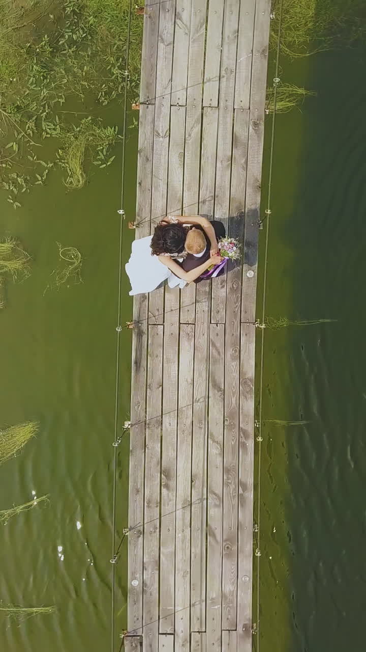 camera turns above happy newly wedded couple standing on old wooden bridge over narrow river in park on sunny day