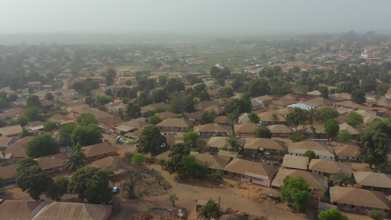 Immagine di una vasta zona residenziale a bassa densità in Guinea-Bissau, Africa occidentale, caratterizzata da tetti marroni e una fitta copertura arborea che si estende verso un orizzonte nebbioso e lontano. Riflette la comunità locale.
