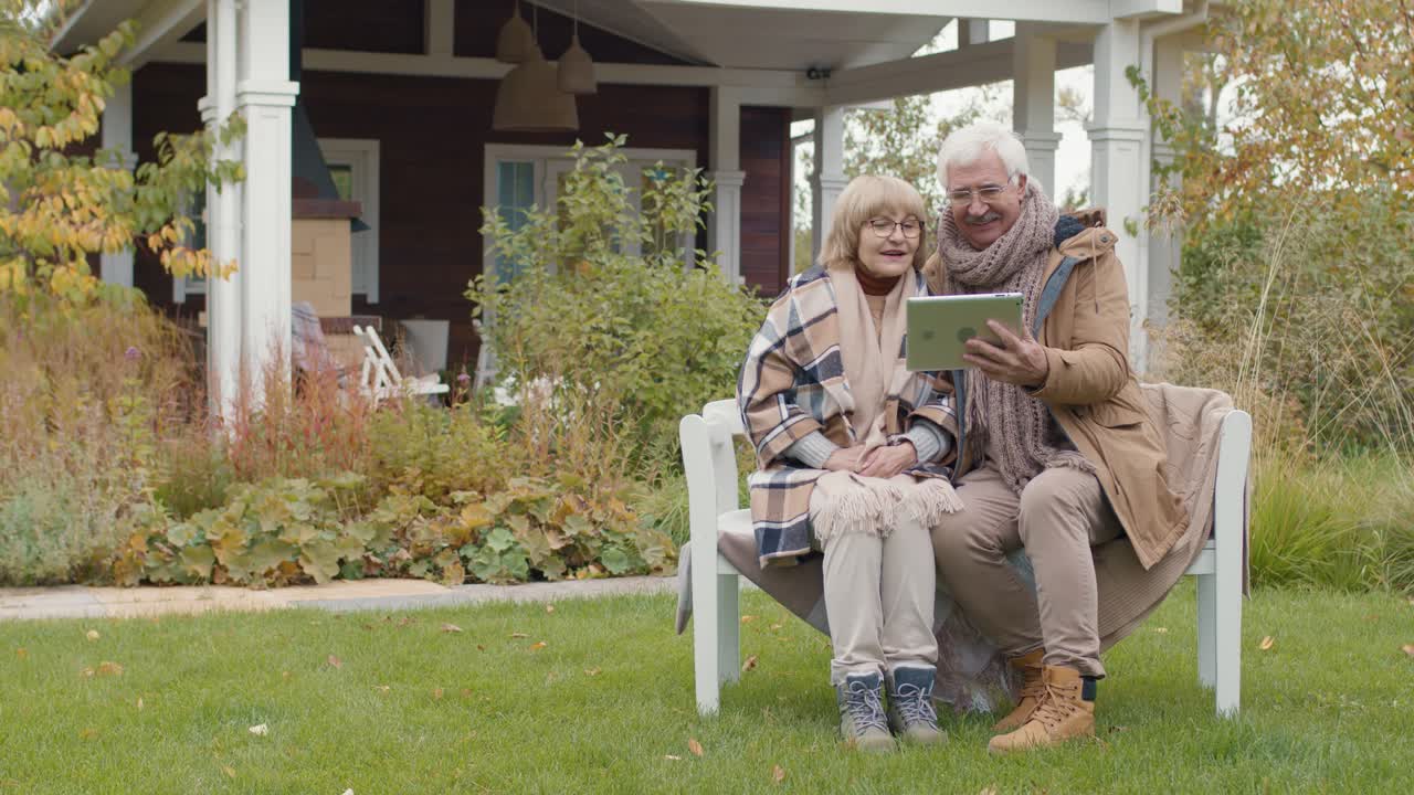 태블릿을 사용하여 화상 통화를 하면서 집 정원의 벤치에 앉아 있는 노부부