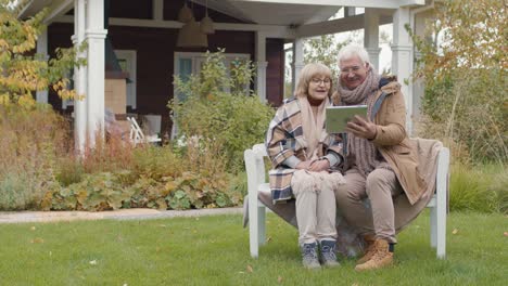 older couple sitting on a bench in their home garden while talking on a video call using a tablet