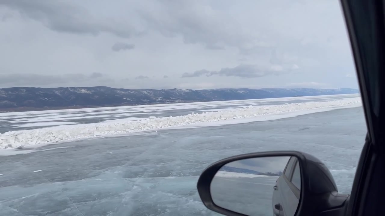 POV da un'auto sulla superficie ghiacciata del Lago Baikal, che mostra una strada di ghiaccio, creste di neve e montagne distanti sotto un cielo coperto in Siberia, Russia