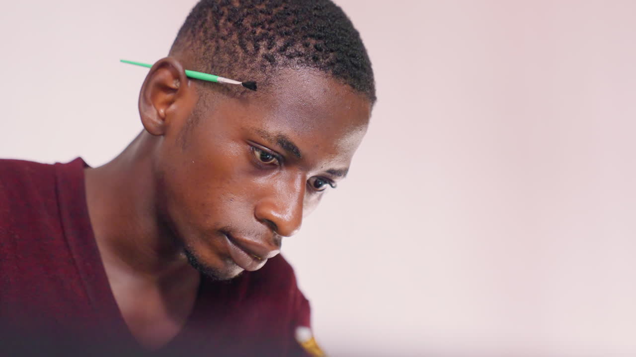 Pintando retrato em close-up de homem negro refletindo com lápis atrás da orelha, luz natural suave, expressão sutil, atmosfera tranquila de ateliê, designer esboçando layout, estudo de conservador, lento
