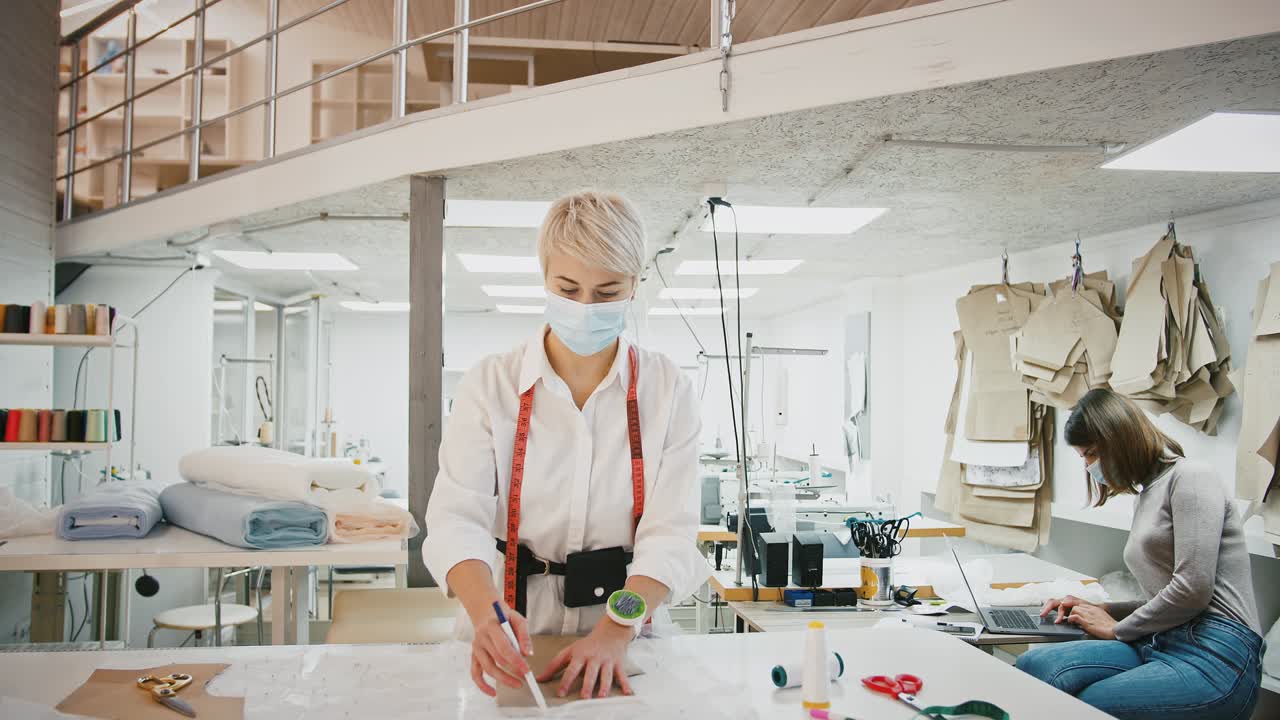Female in protective mask, tailor of sewing workshop is cutting fabric with scissors, colleague working at laptop, coronavirus