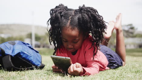 young girl lying on grass using smartphone, enjoying outdoor playtime near backpack