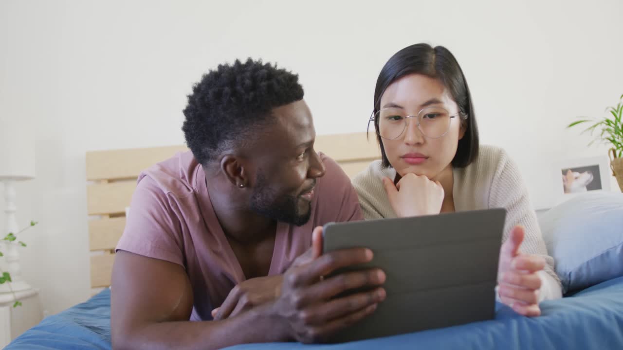 Happy diverse couple using tablet and lying in bedroom