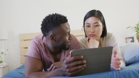 happy diverse couple using tablet and lying in bedroom