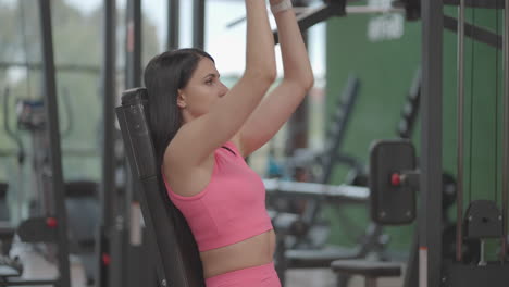 a brunette hispanic woman in a pink suit raises a dumbbell to the top while training her shoulders in the gym. exercise sitting on a bench to train the shoulders and arms. weight training