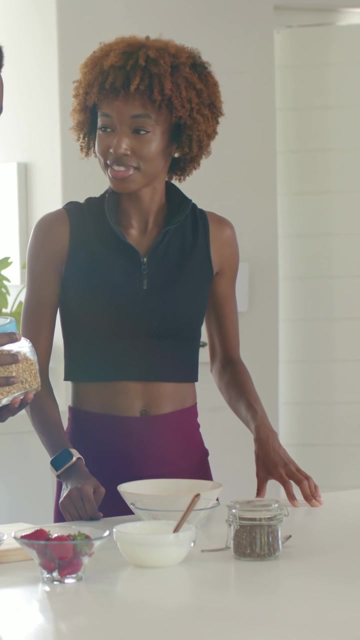 Video verticale: Uomo che porge un barattolo, donna con top da allenamento che controlla la granola al bancone, telefono su treppiede.