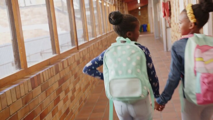 Camera pulling back two girls hand-in-hand going down school hall to exit, packs, pencils drifting