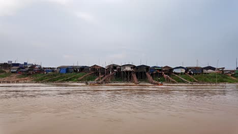 boat view of stilt houses along cambodia's riverbank of chong khneas under a cloudy day.