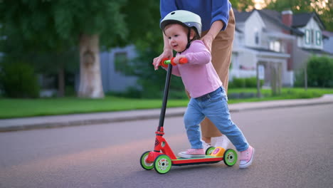 parent helps toddler learn to ride a scooter in the neighborhood