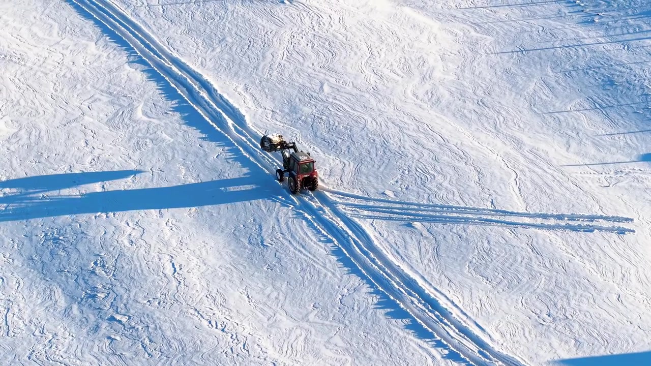 Drone videosu, geniş, el değmemiş karlı bir tarlada ekipman çeken kırmızı bir traktörü, keskin sabah ışığı altında, dokulu kış arazisi üzerine uzun gölgeler düşürerek yakalıyor.