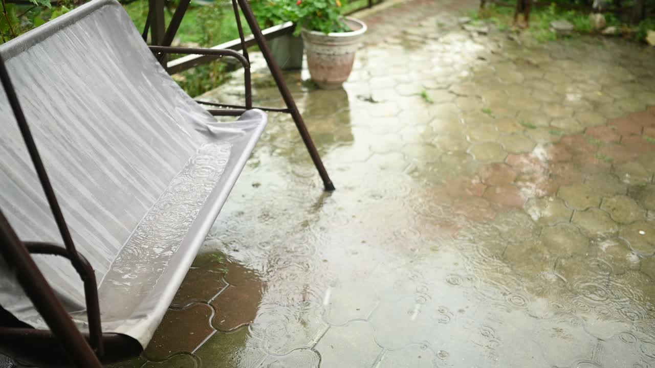Swaying in the wind wet country swing in the country summer house in heavy rain. Circles of raindrops and huge puddles on the asphalt.