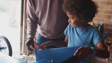 Pre-teen-mixed-race-boy-standing-at-a-workbench-making-a-racing-kart-with-his-father-in-their-garage,-close-up,-waist-up