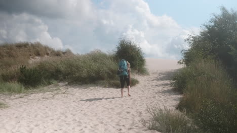 wide shot of a blonde female hiker with heavy backpack walking up a sand dune on a beautiful summer day, råbjerg mile, denmark