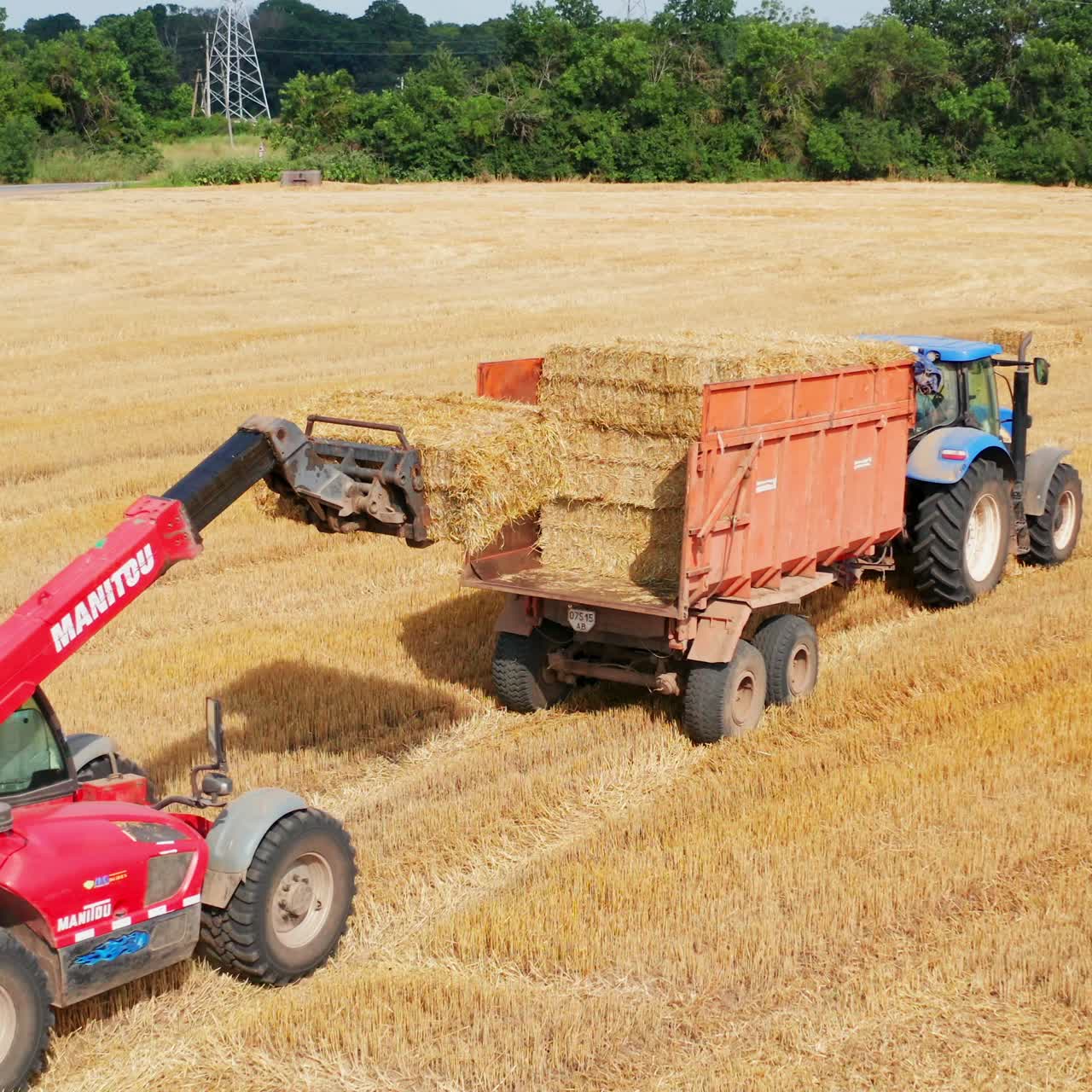 Little excavator picking hay bales and loading them on the tractor. Approaching to the tractor machine full of straw harvest