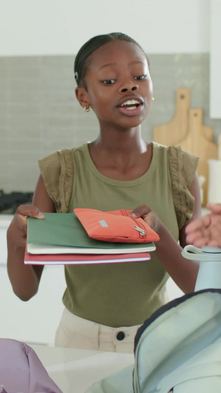 Vertical video: Entering older sister handing books, sisters packing backpack for class in kitchen