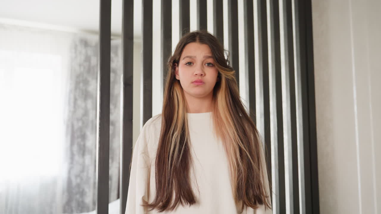Elegant young lady stands before wooden divider as frame widens with gentle zoom out, mouth turns side to side in playful pout, eyes closed, soft daylight on face, calm mood, minimal interior