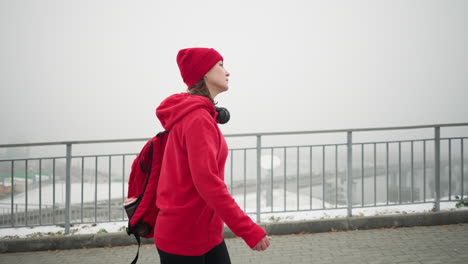 vista lateral de una mujer con gorra roja y chaqueta caminando a lo largo de una barandilla de hierro con auriculares alrededor del cuello y la mochila, paisaje nevado y vista borrosa del puente y el paisaje urbano en una atmósfera de niebla