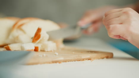 pastry cook cutting bread into bite sized pieces on wooden board with sausage, banana, eggs and bowl on white table under bright morning window light, capturing calm cooking preparation scene