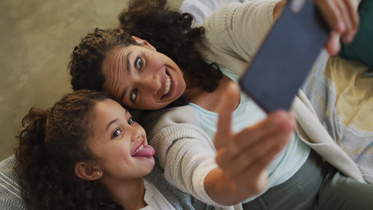 Madre e figlia multietniche felici sdraiate sul pavimento, divertendosi e facendosi un selfie.