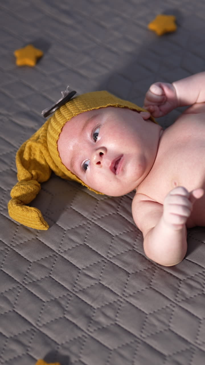 Adorable child in yellow hat and pants lying on the bed face up. Baby in yellow costume with decorative toy stars. View from above. Vertical video
