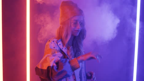 cheerful stylish blonde girl in hat and sunglasses dancing and smiling with the smoke and neon lights