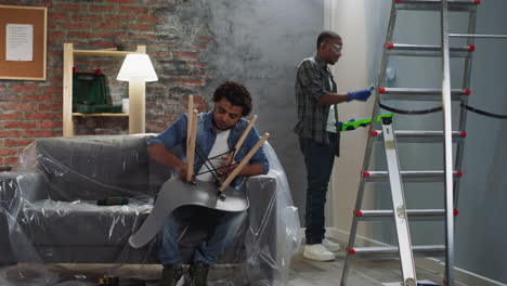 black man paints wall guy assembles chair in living room