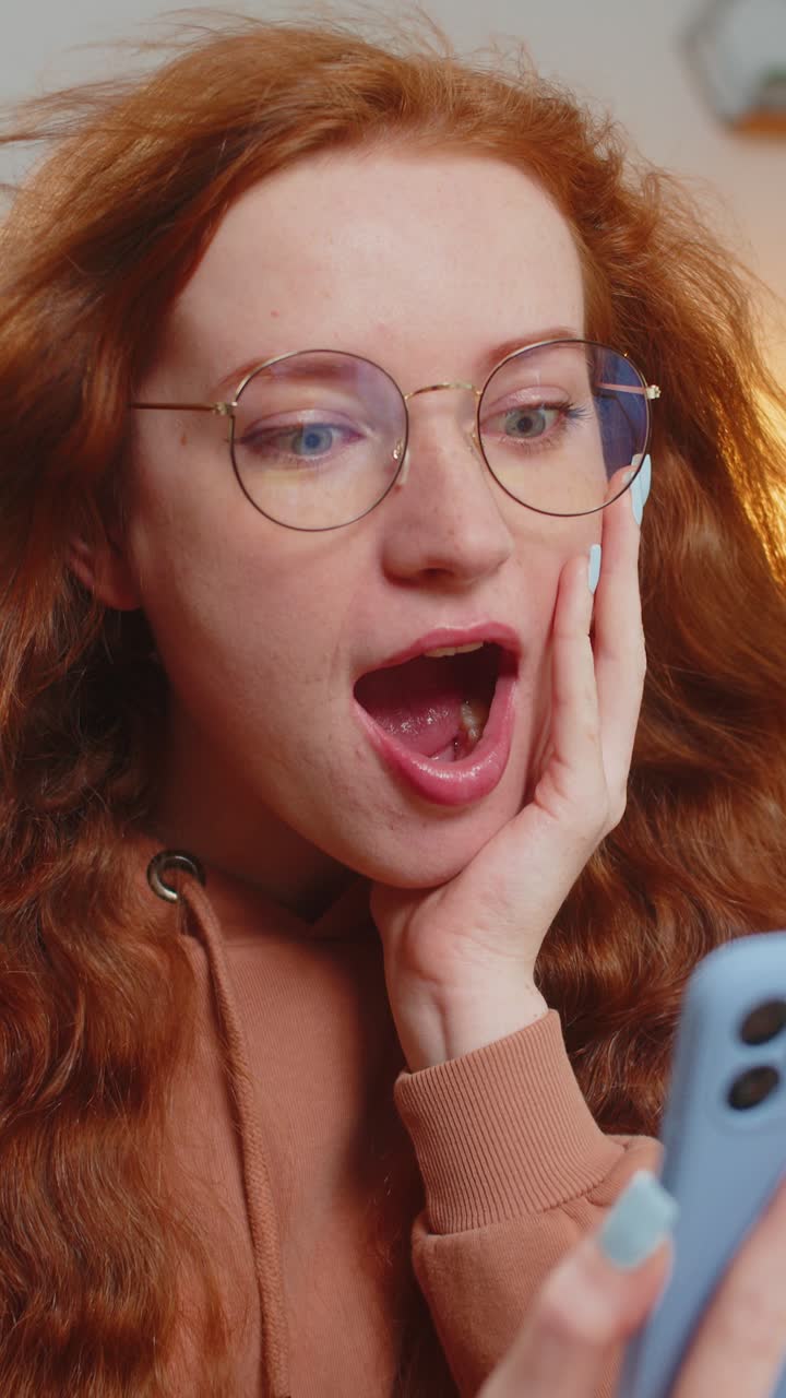 sorprendida mujer caucásica pelirroja con el cabello azotado por el viento usando un teléfono inteligente sentada en el sofá en casa