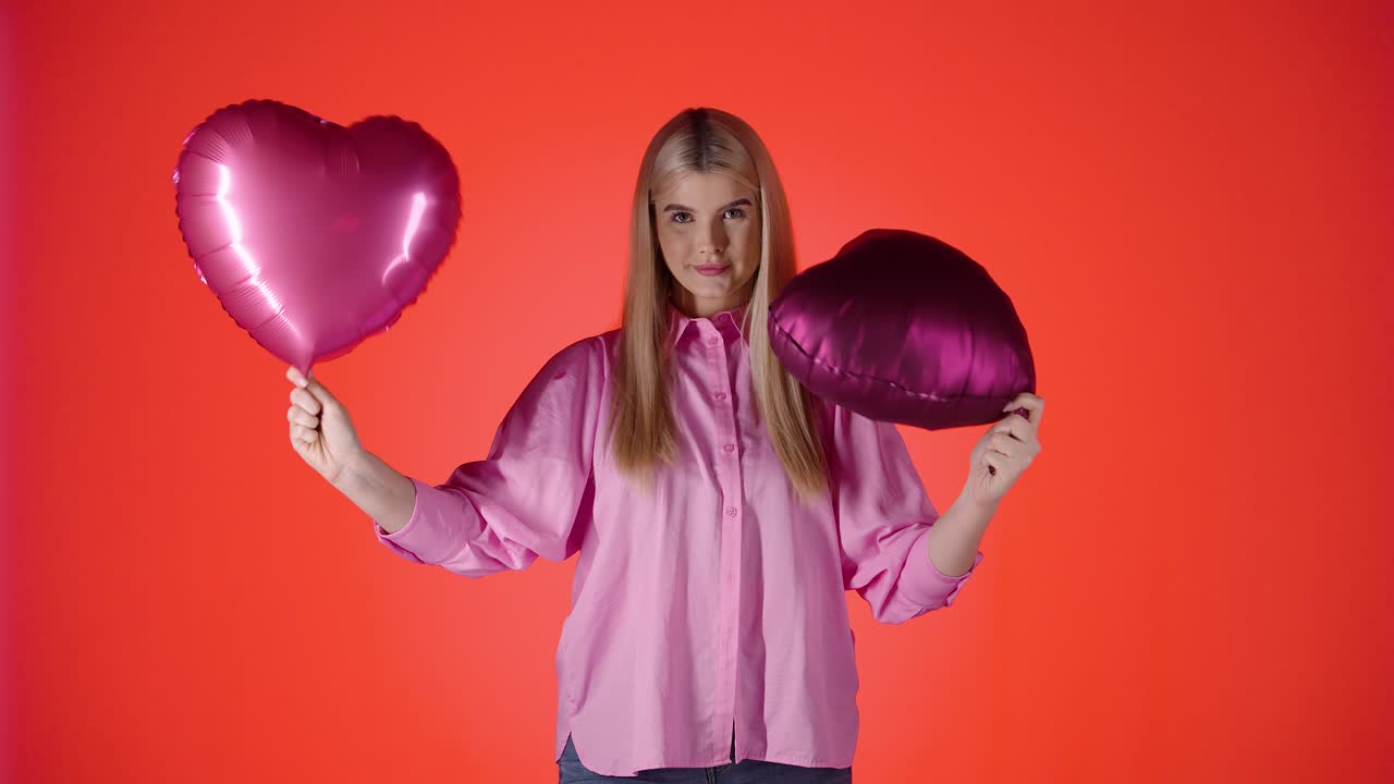 una mujer rubia muy bonita agitando globos en forma de corazón púrpura contra un fondo rojo, una toma de estudio colorida