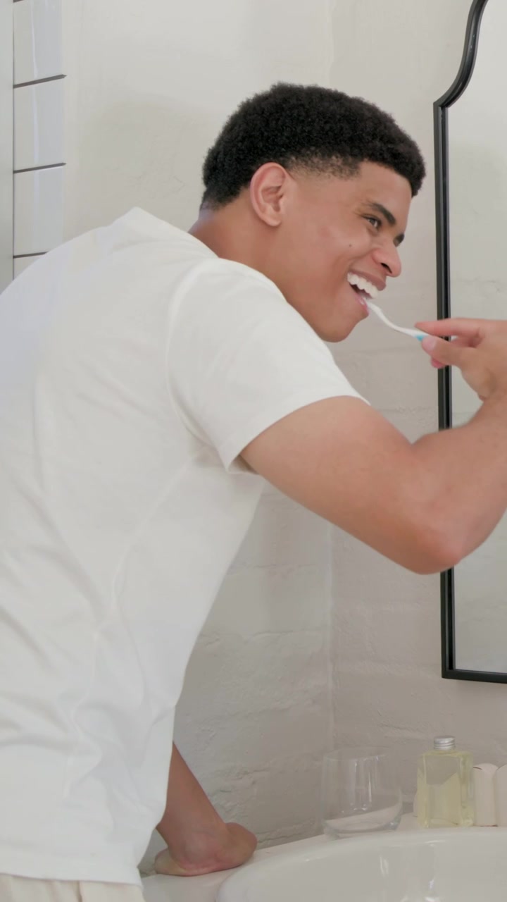 Vertical video: Starting man in white tee brushing teeth at home sink, using toothbrush
