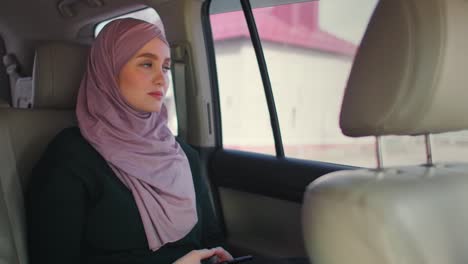 young muslim woman in hijab sitting in car on passenger rear seat. muslim woman day dreaming in back seat. a young woman in a hijab looks out the window.