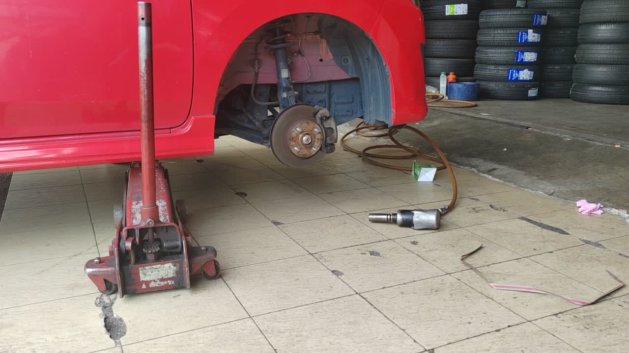 A small red car is seen in a workshop, undergoing a tire change. The front tire is replaced as the car is slightly lifted for ease.