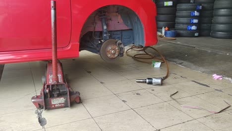 a small red car is seen in a workshop, undergoing a tire change. the front tire is replaced as the car is slightly lifted for ease.