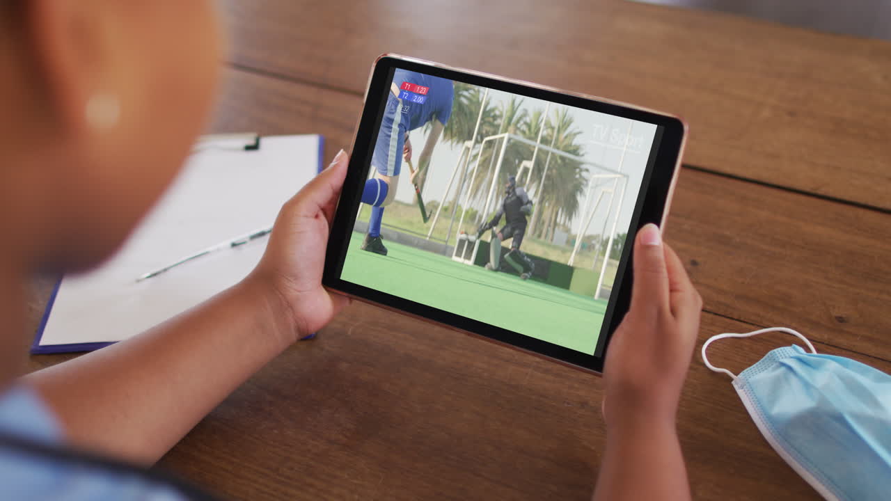 태블릿에서 하키 경기를 시청하는 얼굴 마스크와 함께 테이블에 앉아있는 여성의 복합