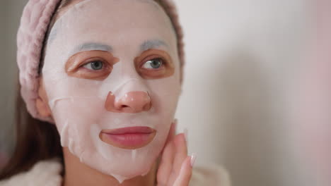 lady smooths cosmetic fabric mask on face with hand. woman in knitted hair band with positive emotions waits for action of nourishing mask in toilet