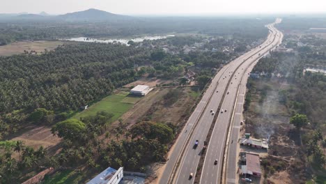 a modern mysuru highway with traffic runs by a large lake, fields, and scattered homes. this aerial shot shows infrastructure development blending with nature under a clear, cloudy sky