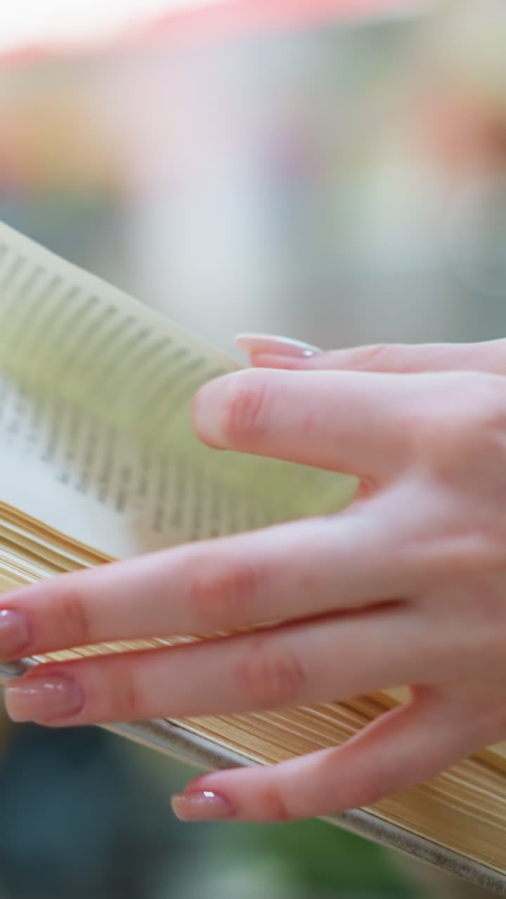 Close up hand view of white woman turning to new page in woven gold sweater, modern structure, blur figures and green decorative flower in background