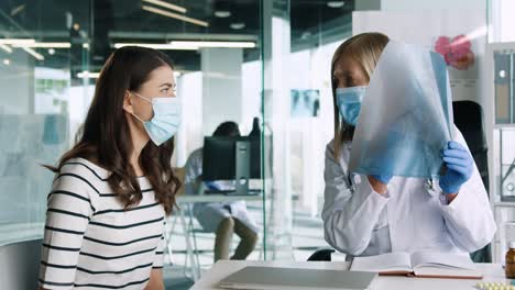 doctora caucásica con máscara médica sentada en el escritorio explicando la enfermedad pulmonar por coronavirus a una paciente en consulta médica