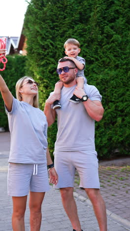 family enjoys outdoor fun with selfie. a family takes a selfie while enjoying a sunny day outdoors, with a child on the father's shoulders and mom posing